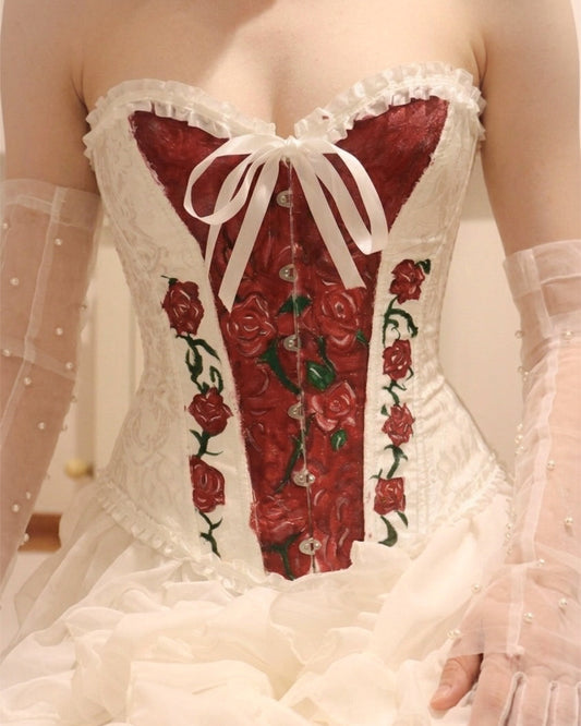 A close-up of a white lace corset with a red rose pattern on the front panel and waist, styled with sheer white gloves.