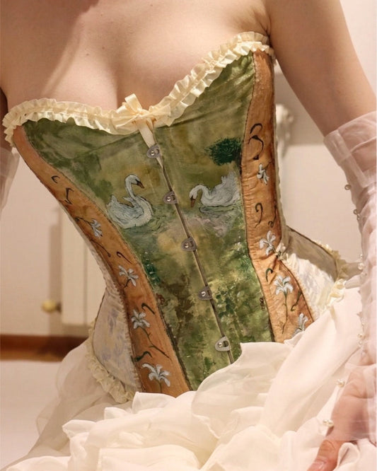 A close-up of a person wearing a Victorian-style corset with a green swan landscape print, gold side panels, cream ruffles, and a white ruffled skirt. Romantic outfit featuring the hand-painted Swan Lake Corset paired with a ruffled white skirt, fairycore and cottagecore aesthetic