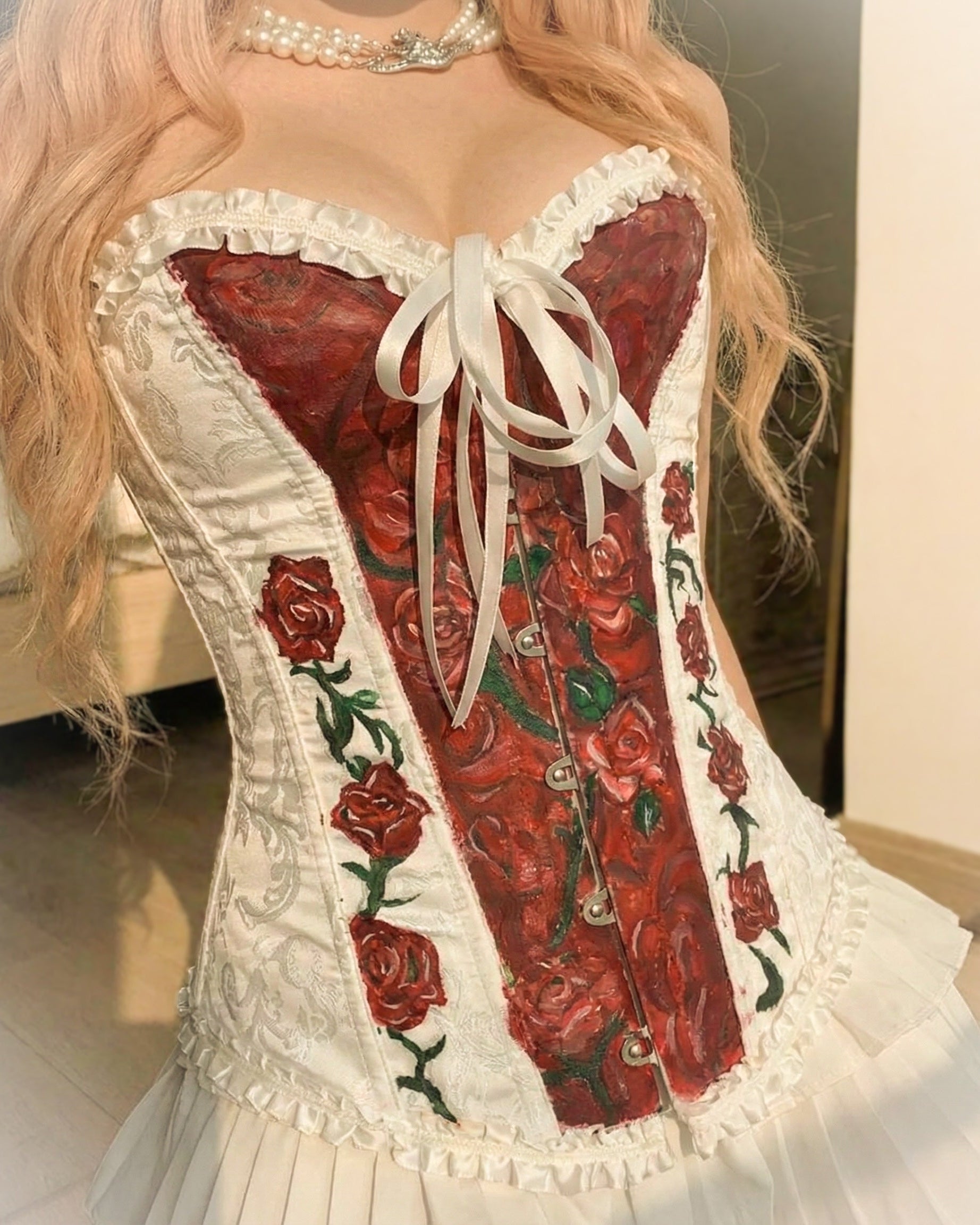 A close-up of a person wearing a Victorian-style corset top with a red rose pattern on the front panels, white ruffled trim, a white bow, and a white ruffled skirt. The person has long blonde hair and is wearing a pearl choker.