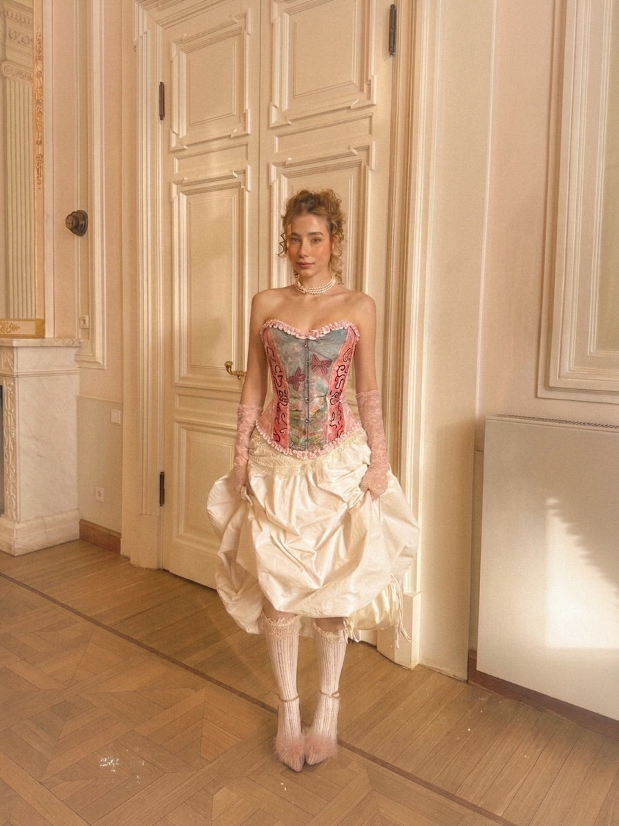 A woman stands in an elegant interior room wearing a Victorian-style corset dress with a pastel multicolor corset bodice, sheer pink long gloves, and a voluminous cream skirt.