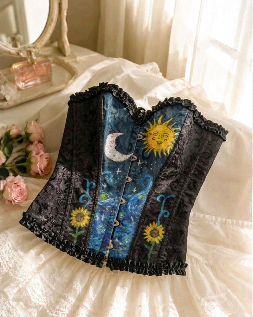 A gothic-style corset top with a dark lace-trimmed sweetheart neckline, featuring a celestial print of a yellow sun, crescent moon, and blue stars on a dark background, styled on a white lace bed with pink roses.