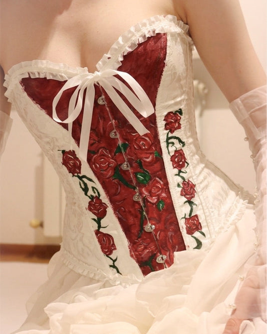 A close-up of a white and red corset-style dress with embroidered red roses, lace trim, and a white bow at the bust, worn by a model in a softly lit indoor setting.