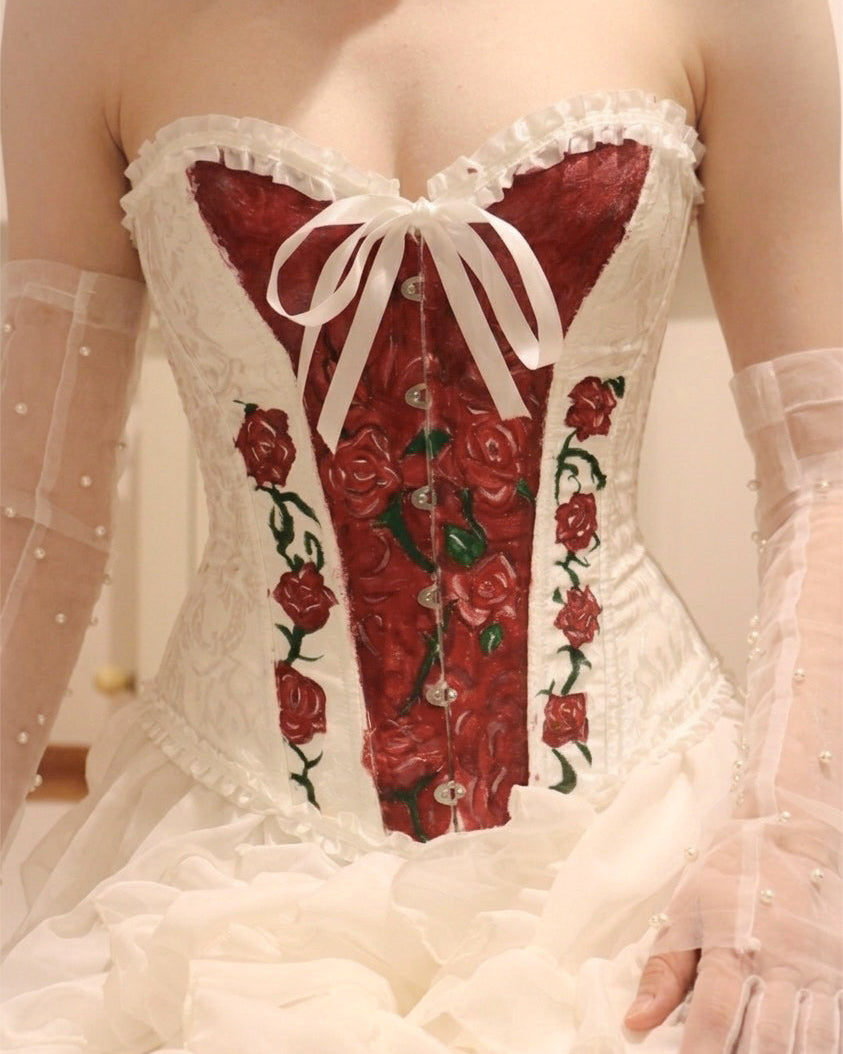 A close-up of a white lace corset with a red rose pattern on the front panel and waist, styled with sheer white gloves.