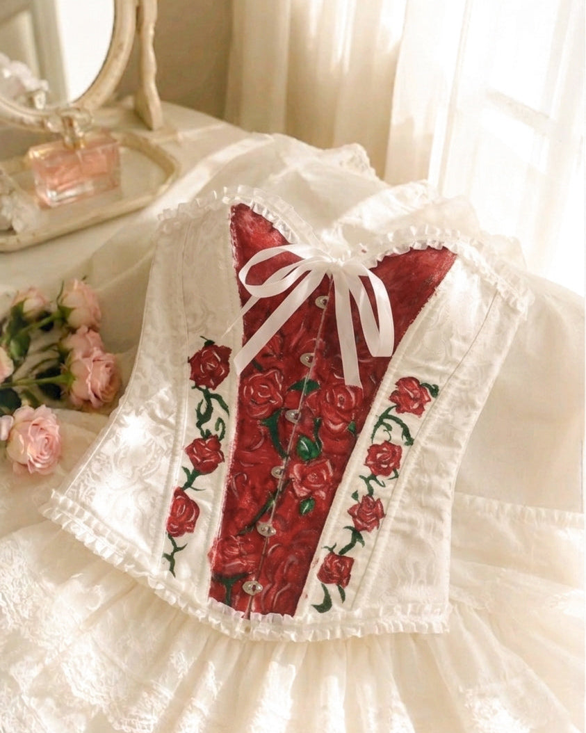 A white lace corset-style top with a red floral embroidered center panel, red rose embroidery along the sides, and a white bow at the neckline, displayed on a white ruffled skirt against a softly lit indoor background.