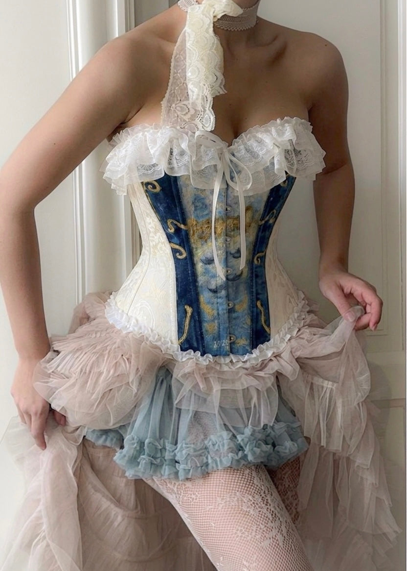 A model wearing a Victorian-style corset dress with a blue and gold patterned corset, white lace trim, and a layered tulle skirt in pale pink and light blue.