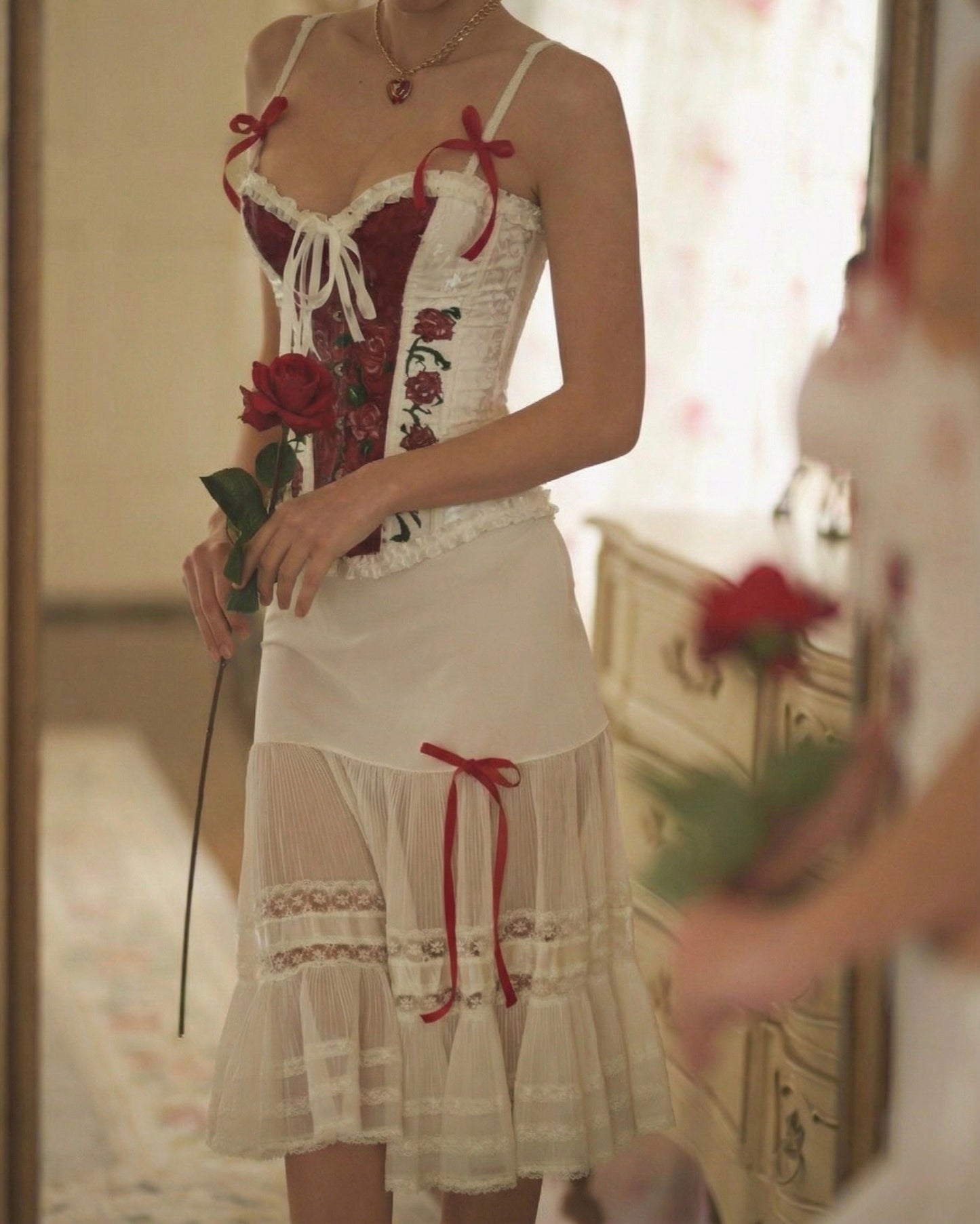 A woman wearing a white and red lace corset-style lingerie dress with red rose embroidery, holding a red rose in a softly lit indoor room.