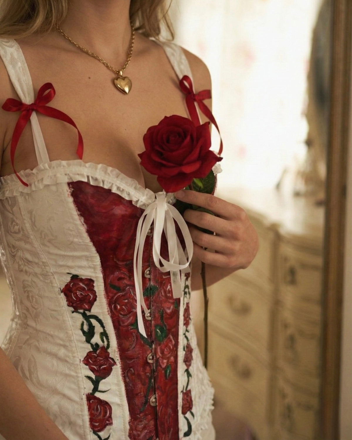 A close-up of a woman wearing a white corset-style lingerie top with red rose embroidery and red ribbon ties, holding a red rose in a softly lit indoor setting.